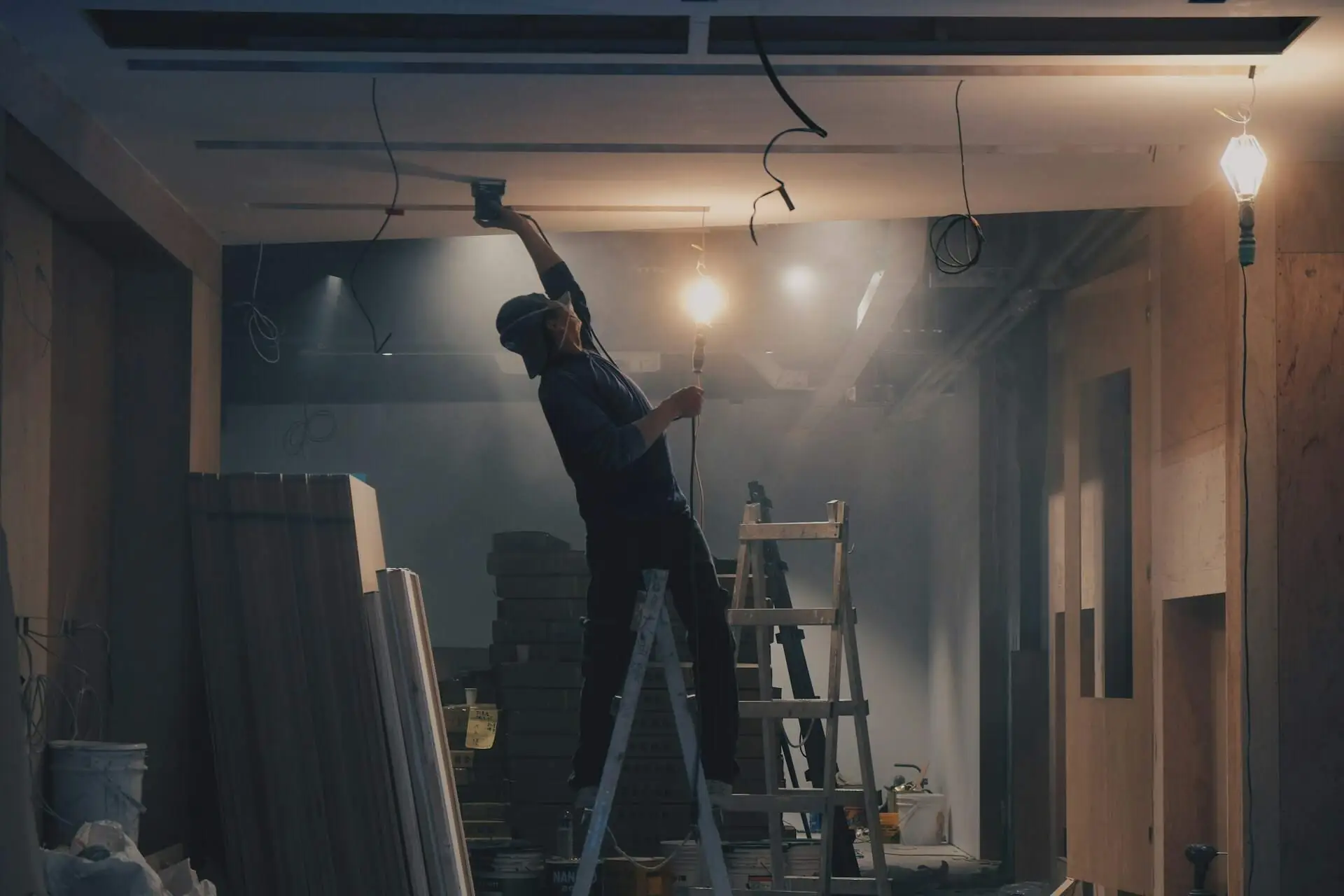 A painter on a ladder sanding a ceiling under work lights before painting